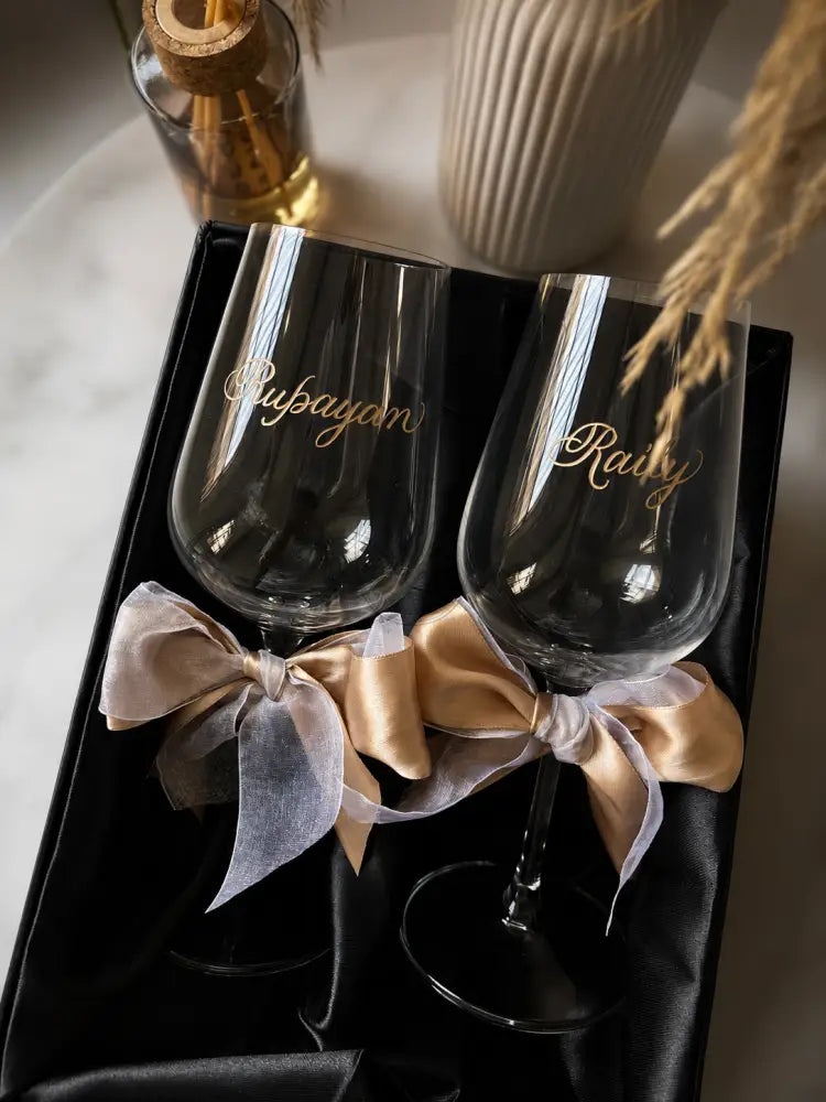 Two black glasses with gold text and ribbons in a box, with a cork and bottle in the background.